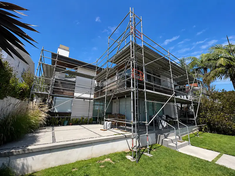 Frame scaffold set up around a modern two-story home with a palm tree and landscaped yard